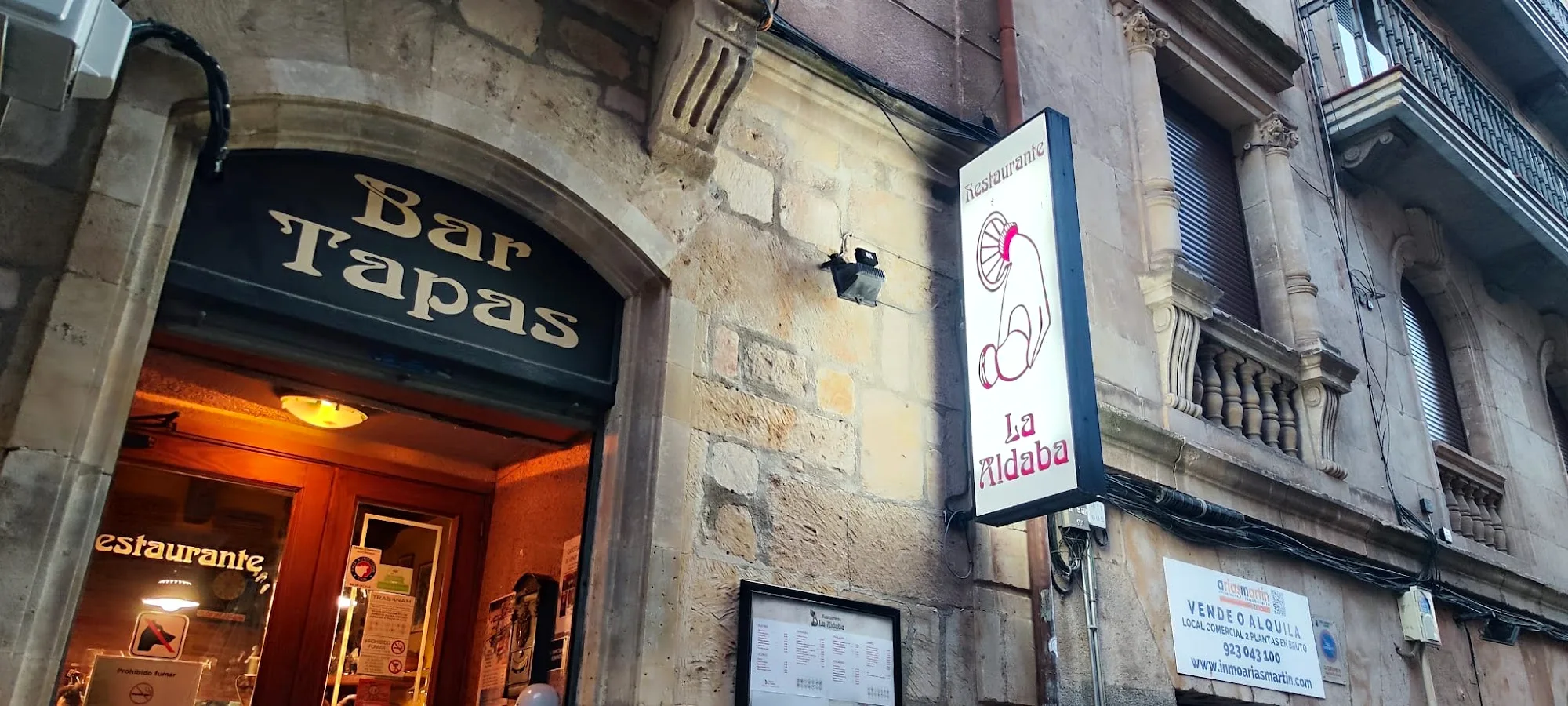 Fachada de piedra de La Aldaba, con el arco de la entrada, el rótulo de Bar Tapas y el cartel luminoso del restaurante